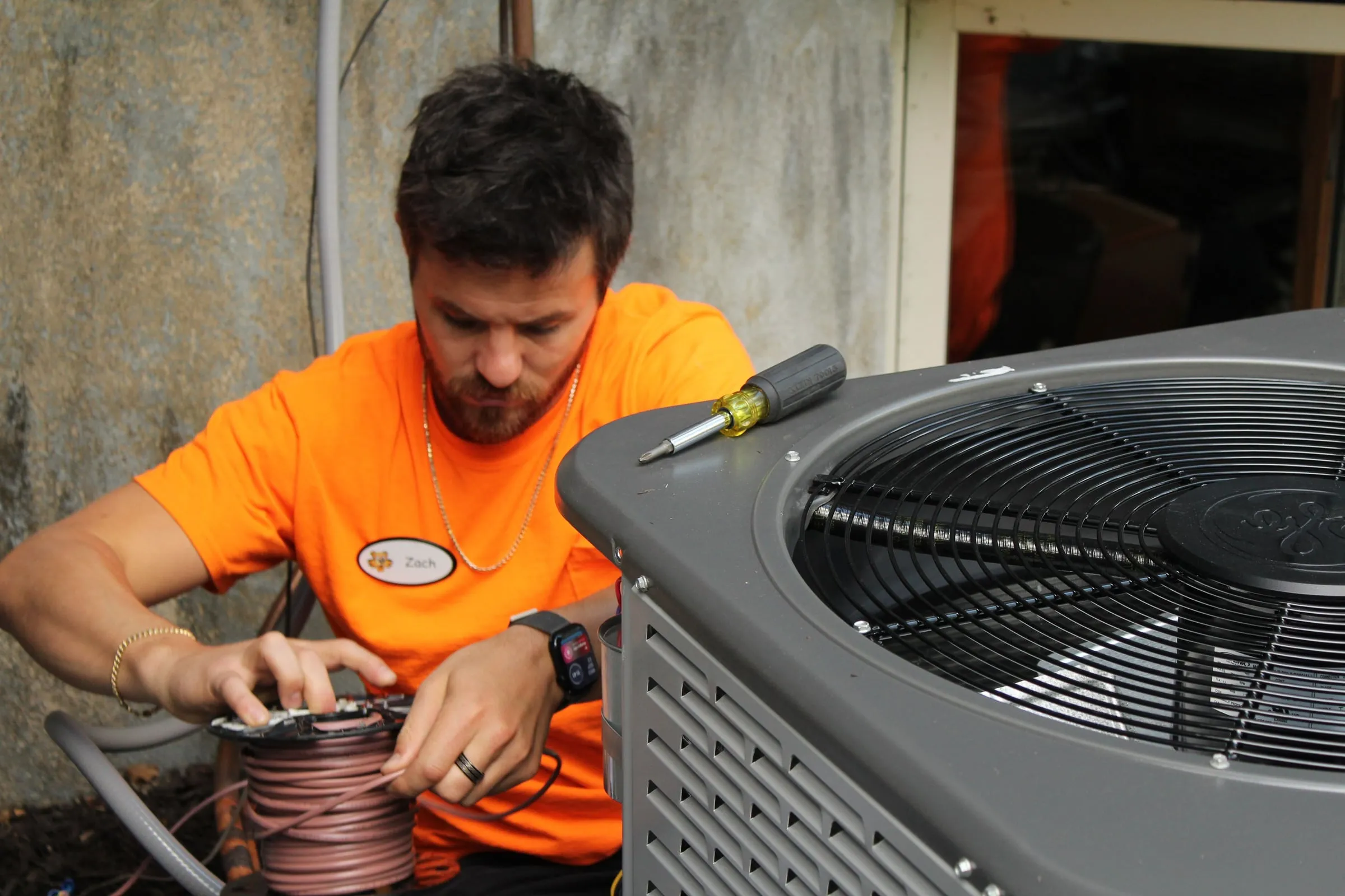 Technician crouching beside a gray outdoor condenser, pulling thermostat wire from a spool